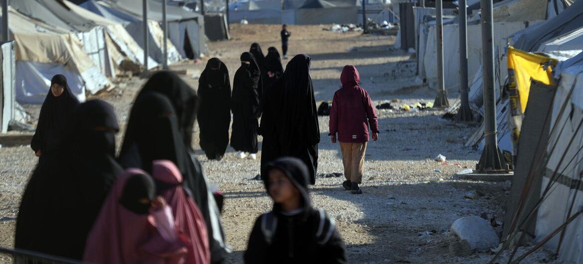 Women and children walk through the Al-Hol Camp in Syria, a displacement site housing over 30,000 people, with tents and infrastructure visible under a clear sky.