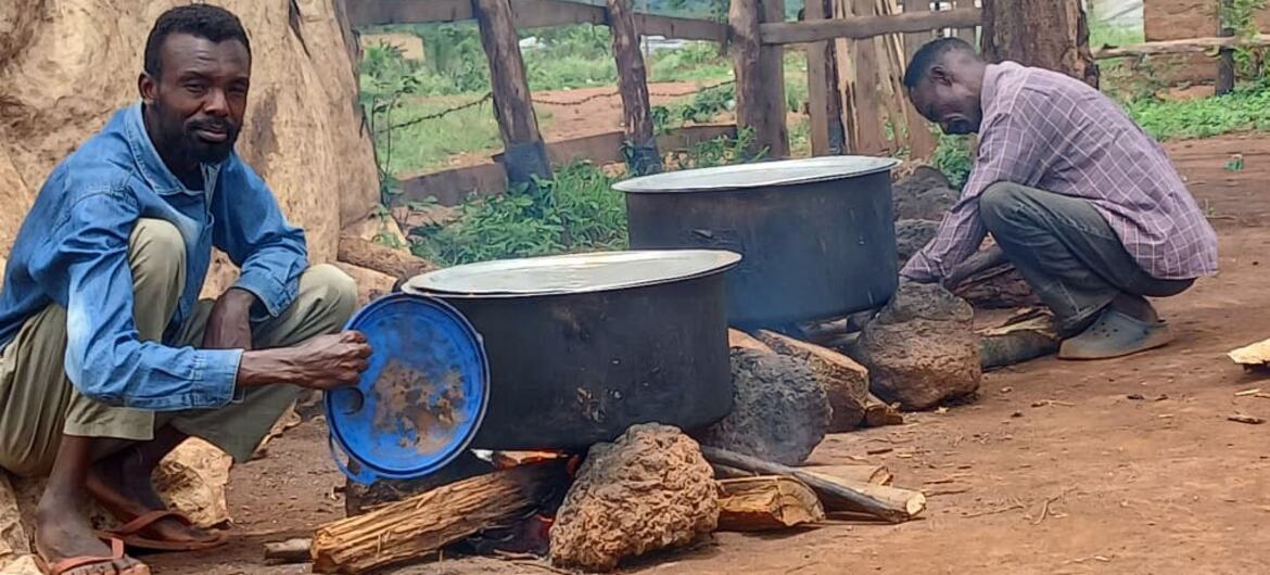 Two African men cooking outdoors in Uganda or Darfur. One man in a blue shirt crouches by a large pot over an open fire, while another man in a plaid shirt tends to a second pot.