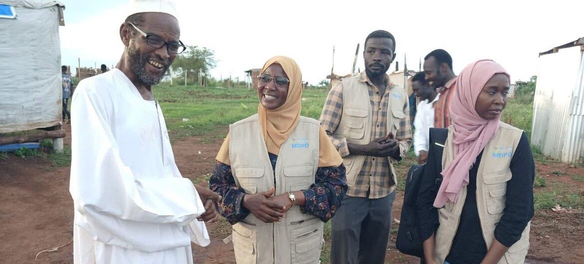 An African man in white robes and a cap shakes hands with two female aid workers wearing MDPD vests and hijabs in a rural outdoor setting.