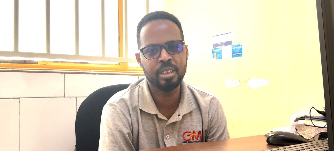 A portrait of a black man with glasses and a beard, wearing a grey polo shirt with a 'GM' logo, sitting at a wooden desk in an office.