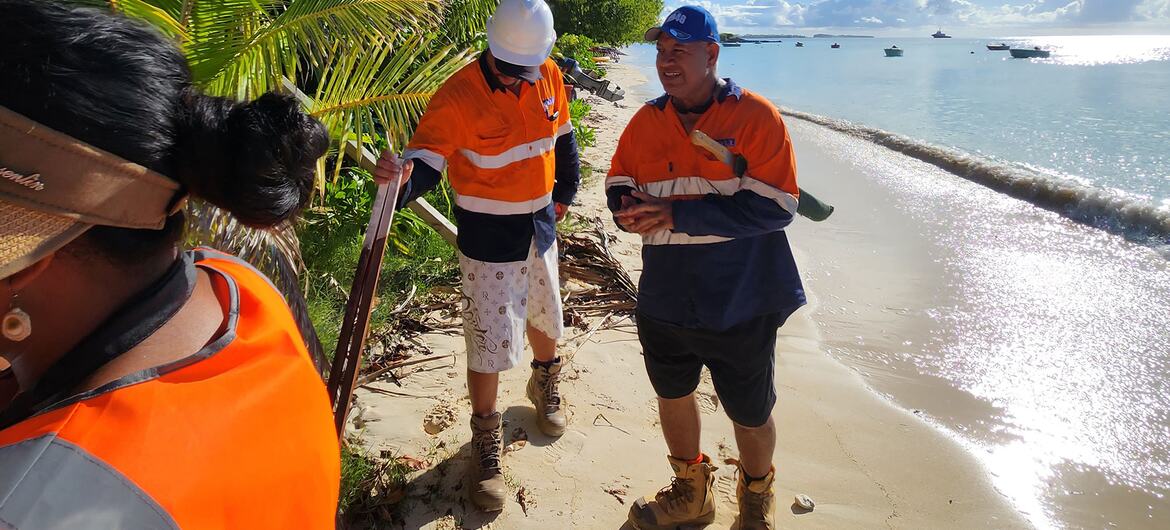 Construction workers in high-visibility orange vests and hard hats on a sandy beach with coconut palms, preparing for coastal protection works on Funafuti, Tuvalu.