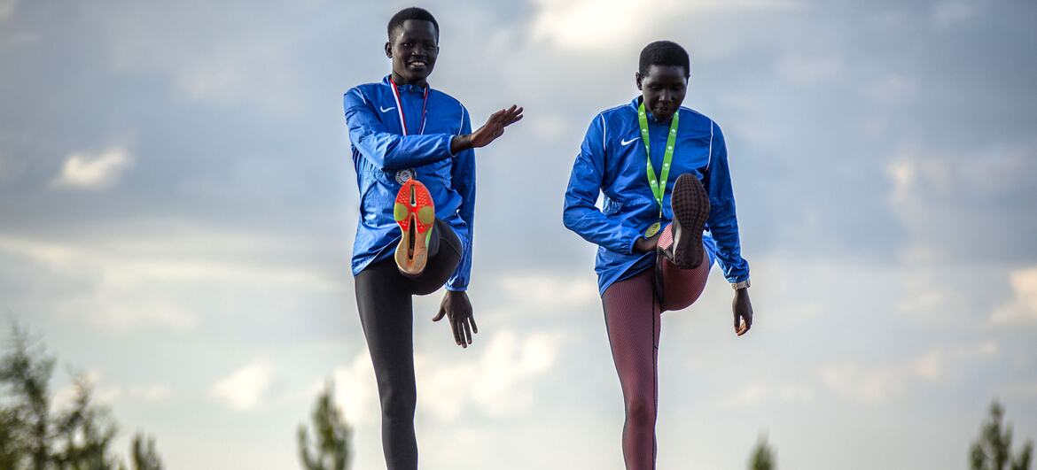 Two young African female athletes in blue tracksuits stretch on a running track at the Teryet National High Altitude Training Centre in Uganda. The image is part of a UNFPA documentation effort to highlight how athletics provides safe spaces and builds confidence for girls, particularly survivors of Female Genital Mutilation (FGM).