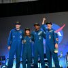 CSA (Canadian Space Agency) astronaut Jeremy Hansen and NASA astronauts Reid Wiseman, Victor Glover, and Christina Koch smile at the crowd during a news conference. They are all wearing blue jumpsuits with patches on the arms and chest areas. Wiseman raises his right fist in a victorious pose.