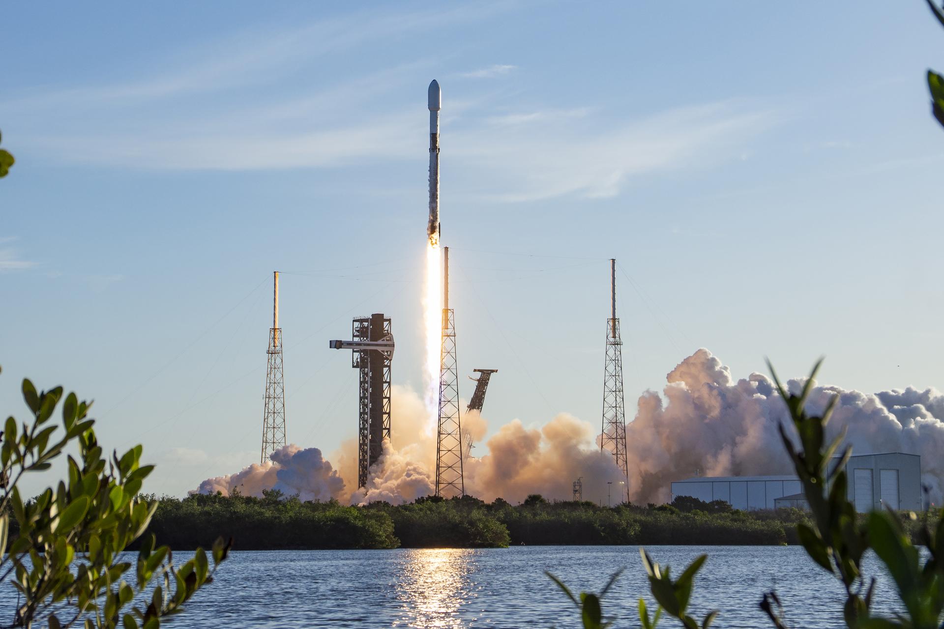 Image shows a rocket launching vertically in a morning blue sky with white clouds at Cape Canaveral Space Force Station in Florida on Saturday, April 11, 2026. The launch reflects in a nearby body of water and shrubs. Photo credit: SpaceX