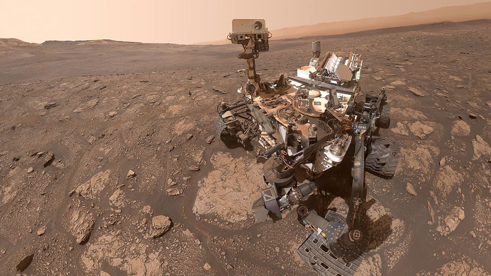 The six-wheeled Curiosity rover is positioned in the right side of the frame on a rugged, rock-strewn, brownish-red Martian plain. The rover’s articulated robotic arm and camera mast are raised. Distant hills and a hazy, dusty orange-brown sky are in the background.
