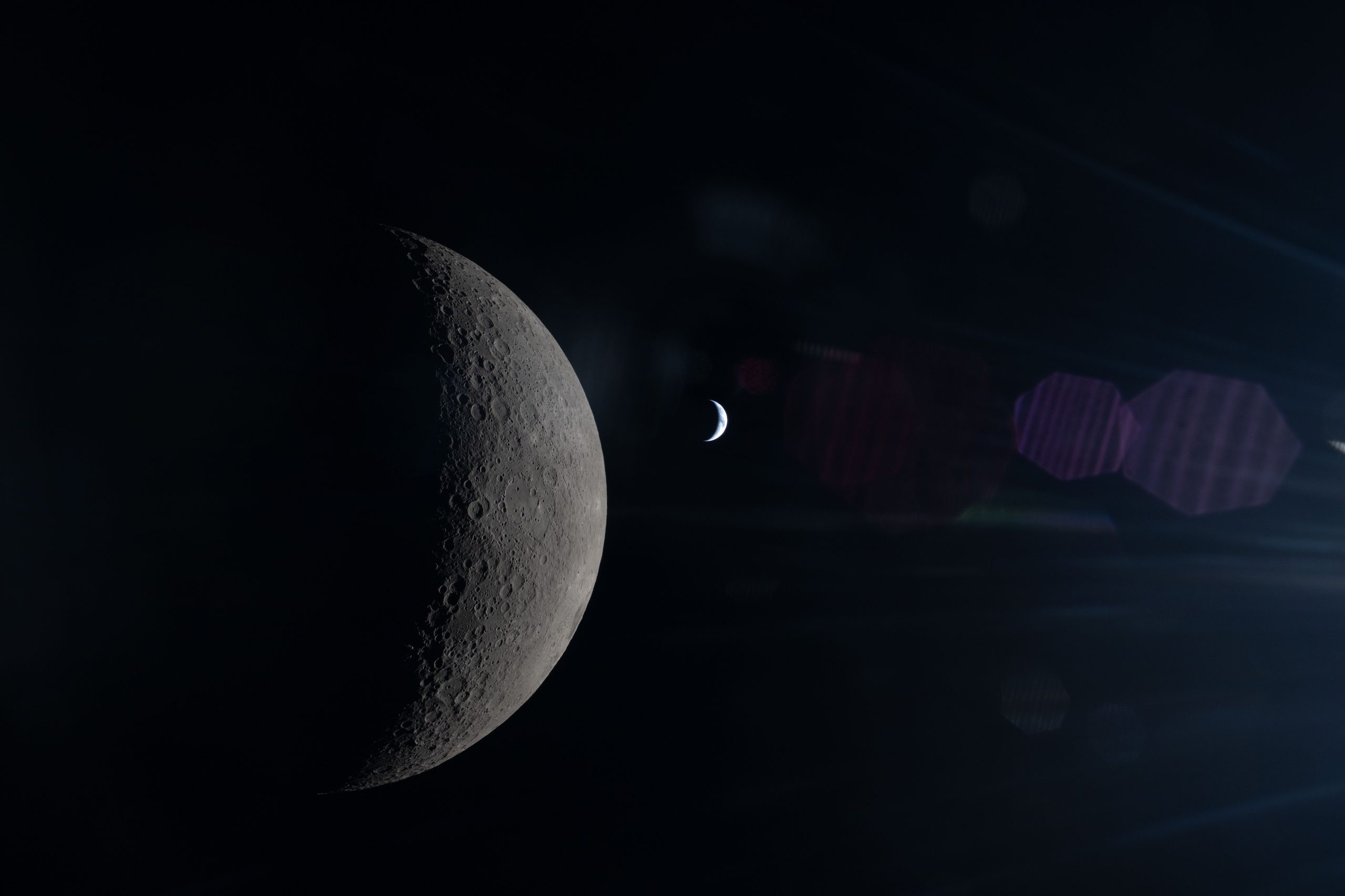 This image, taken from behind the Moon, shows its half-illuminated surface in sharp detail. To the right, a much smaller crescent Earth can be seen, with shades of blue and white just barely distinguishable. A faint reflection of the Orion spacecraft's window overlays the scene, especially in its right half.