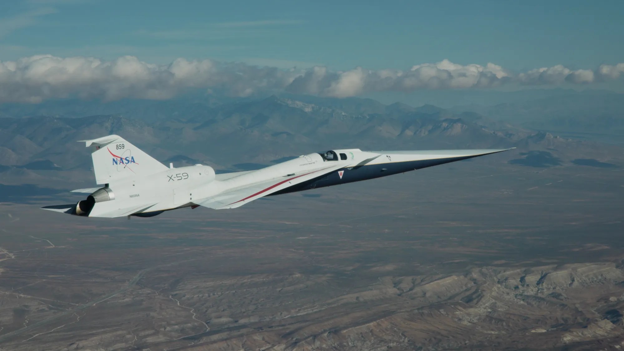 NASA’s X-59 flies above the Mojave Desert on a clear day. The white aircraft has light gray, red, and blue accents, with a NASA logo and the number 859 on its tail. It appears flying level over the desert landscape, with a mountain range visible on the horizon and a trail of clouds above.