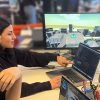 Two students sit at a table with a laptop and large monitor that displays a simulation of a drone flying in an urban environment.
