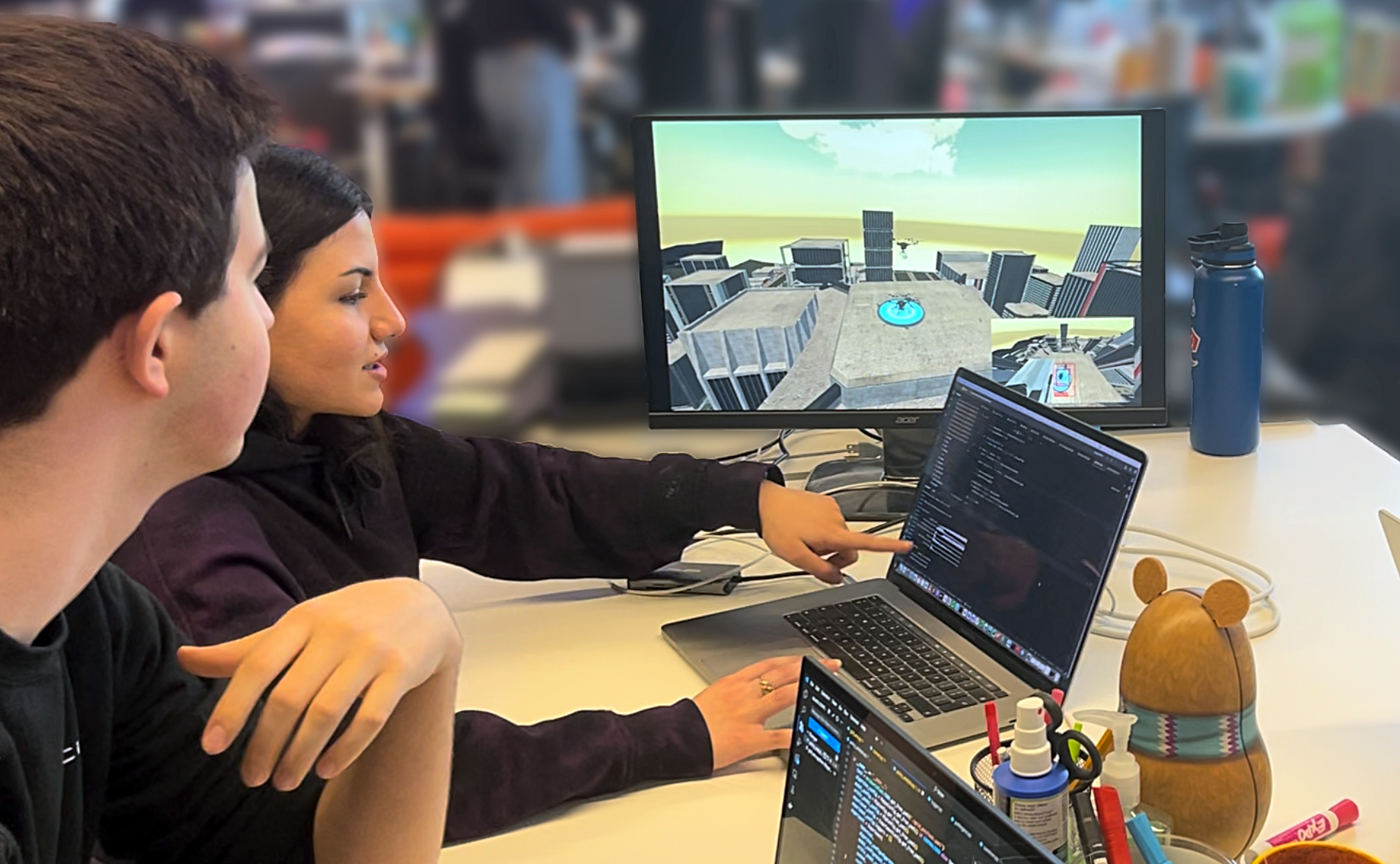 Two students sit at a table with a laptop and large monitor that displays a simulation of a drone flying in an urban environment.