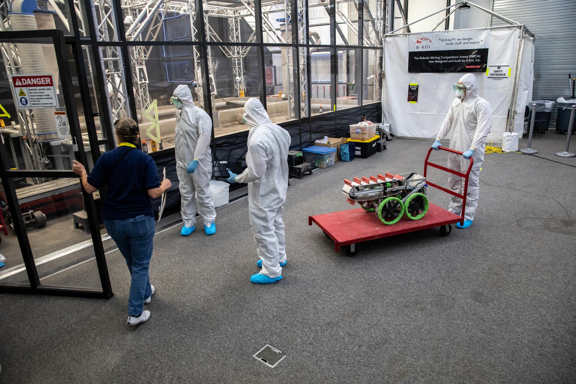 Image shows three people in white protective suits inside a room with another person wearing a blue shirt and blue jeans at NASA's Kennedy Space Center in Florida in 2022. Around them is a red cart with a silver structure and green looking wheels. Photo credit: NASA/Kim Shiflett